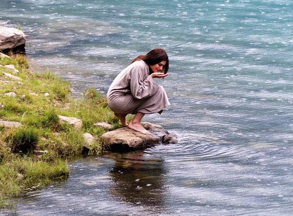 Eine Frau hockt auf einem Stein am Ufer und schöpft mit den Händen Wasser aus dem See.
