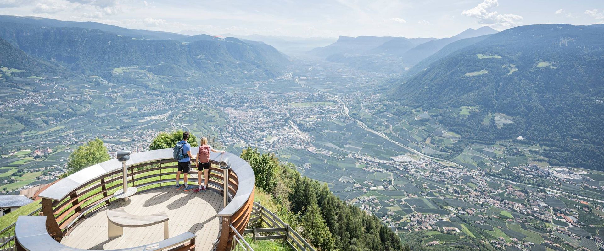Zwei Personen stehen auf einer runden Aussichtsplattform über dem Tal mit Blick auf Berge und Orte in Südtirol.