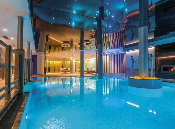 A large indoor thermal spa pool with bright blue water, columns, lounge seating and reflected ceiling lights.