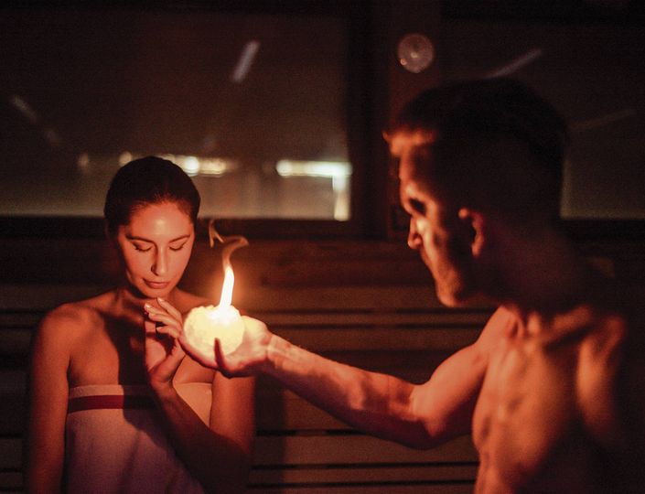 A man holds a candle close to a woman inside a dim sauna.