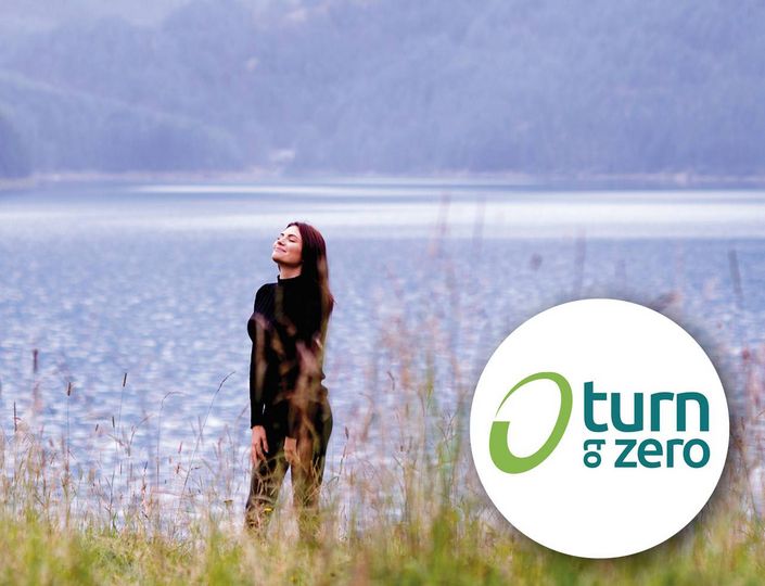Eine Frau steht am Seeufer vor ruhigem Wasser und Bergen; rechts ist ein rundes Turn-to-Zero-Logo zu sehen.