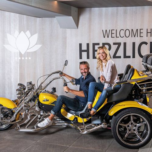 Ein Mann und eine Frau sitzen auf einem gelben Trike in einem Innenraum mit Willkommenswand.