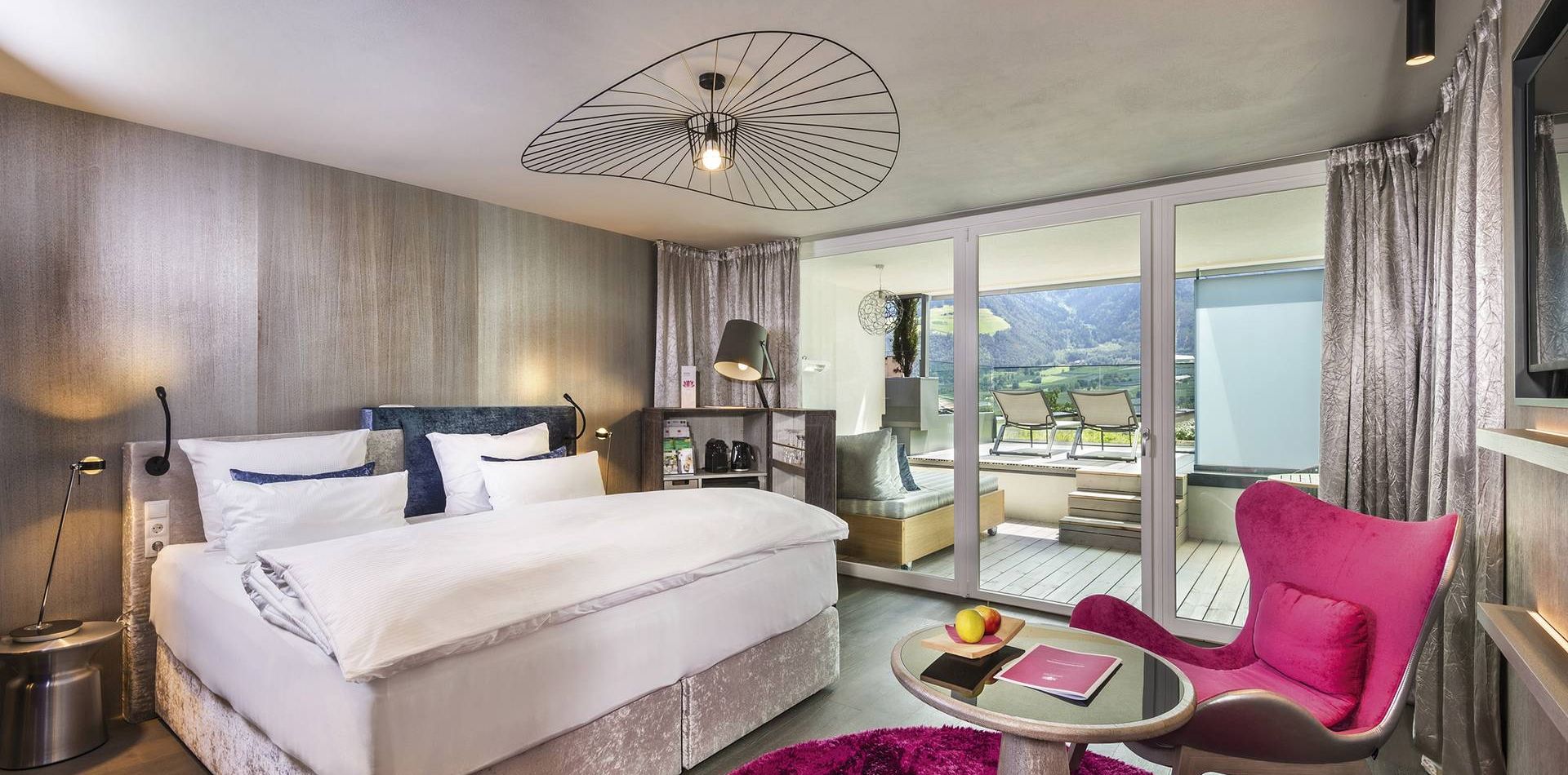 A modern double room with a large bed, pink chair, small table and balcony doors overlooking the mountains.