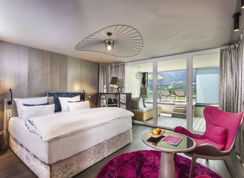 Comfort Room Romantic Fire at Preidlhof A modern double room with a large bed, pink chair, small table and balcony doors overlooking the mountains.