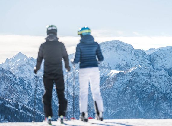 Zwei Skifahrer stehen auf einer Piste und blicken auf die verschneiten Berge.