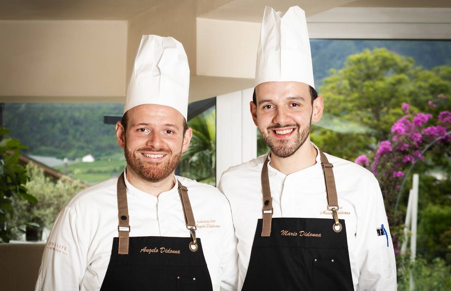 Köche im Gourmetrestaurant des Preidlhof Zwei Köche stehen in der Küche vor einem großen Fenster mit Blick ins Grüne und in die Berge.