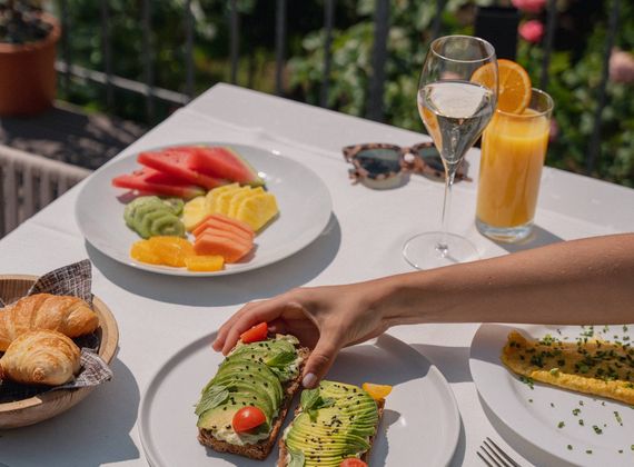 Auf dem Tisch stehen frisches Obst, Croissants, Avocado-Toast, Saft und ein Glas Sekt im Freien.
