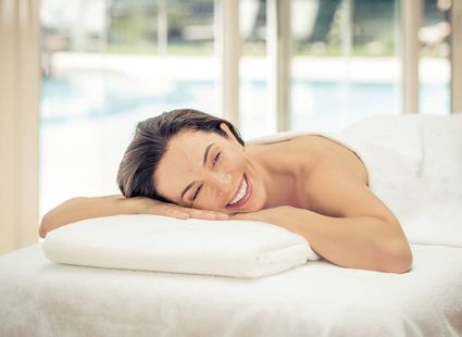 Una donna sorridente è sdraiata su un lettino da massaggio con la testa appoggiata su un cuscino bianco in una sala luminosa.