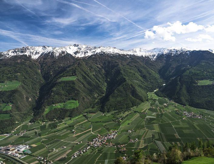 A wide alpine valley with patchwork fields, scattered villages and snow-covered mountains under a blue sky.