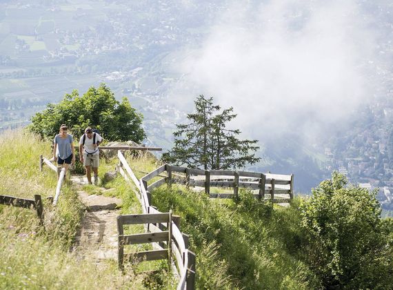 Zwei Wanderer steigen auf einem schmalen Weg mit Holzgeländer in der Berglandschaft auf, im Hintergrund liegt das Tal im Dunst.