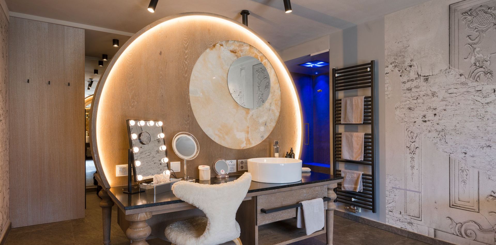 Ein beleuchteter Schminktisch mit Spiegel, Stuhl und Waschbecken in der Penthouse Suite DolceVita im Preidlhof.