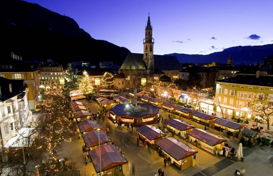 Weihnachtsmarkt mit Kirche in Naturns Der Weihnachtsmarkt ist abends mit beleuchteten Ständen und einer Kirche im Hintergrund zu sehen.