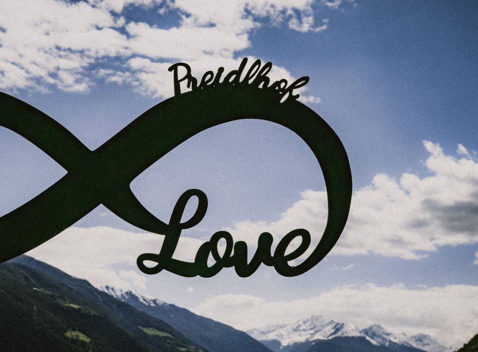 Die schwarze Love-Skulptur mit Preidlhof-Schriftzug steht vor Himmel und Bergen.