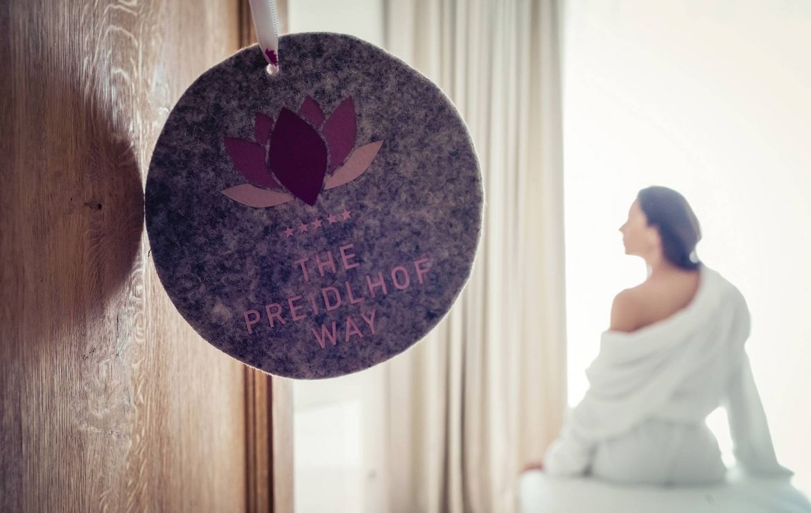A woman sits wrapped in a white robe beside a hanging sign in a softly lit spa treatment room.