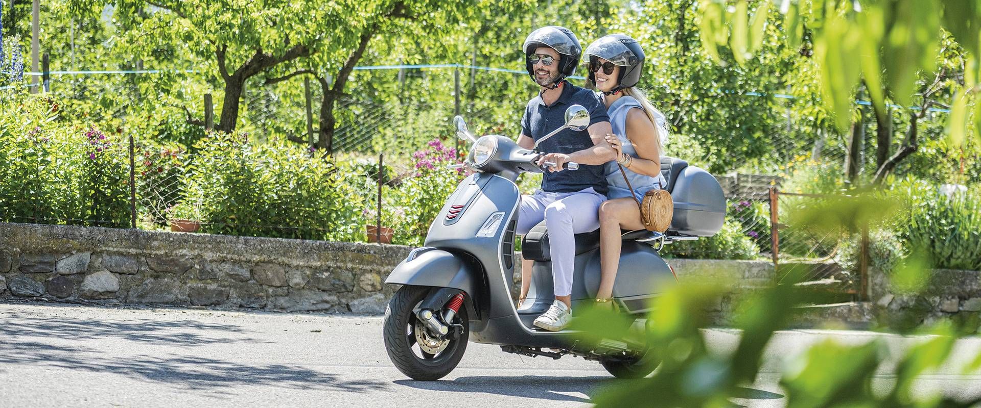 Zwei Personen fahren gemeinsam auf einer Vespa an einer grünen Steinmauer entlang.