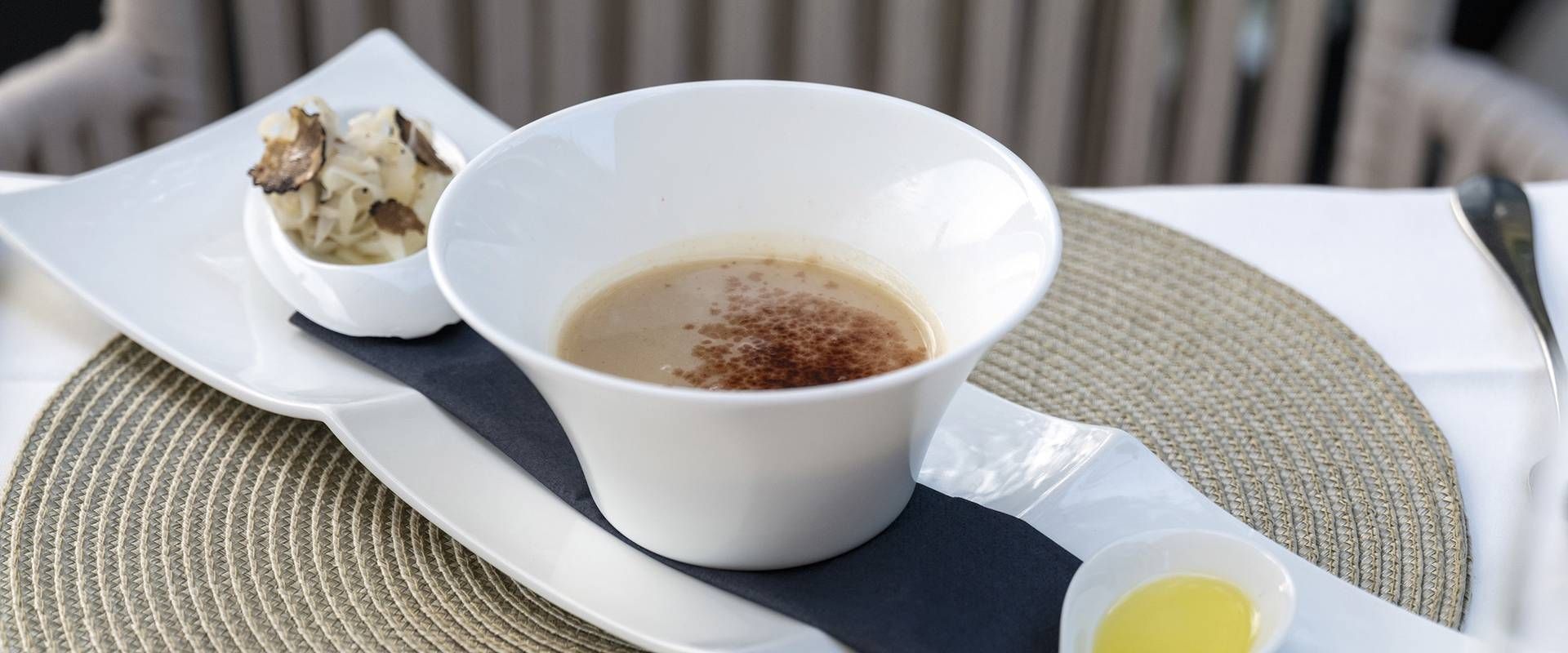 A white cup of coffee sits on a breakfast setting with a small pastry and butter on a woven placemat.