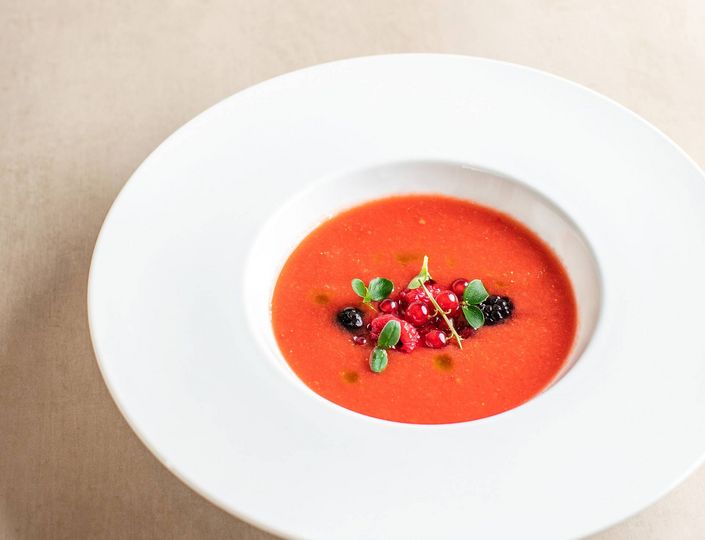 A white bowl holds smooth tomato soup topped with herbs and berries.