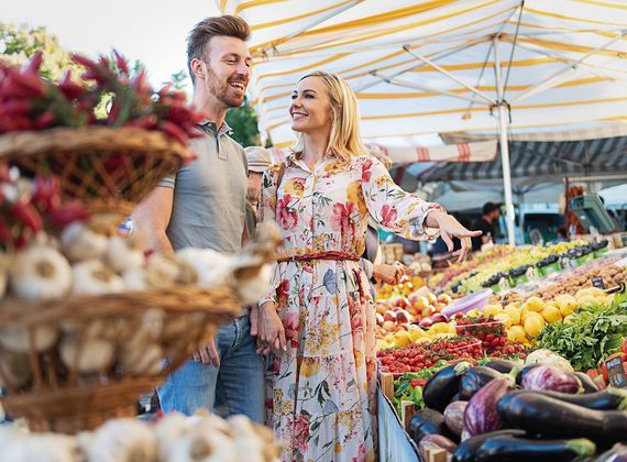 Ein Paar steht am Obst- und Gemüsemarkt und lächelt zwischen Körben und farbig ausgelegten Waren.