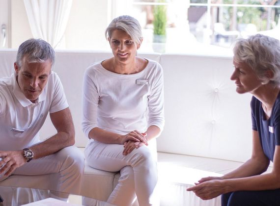 Three people sit together in a bright lounge, discussing papers during a medical consultation.