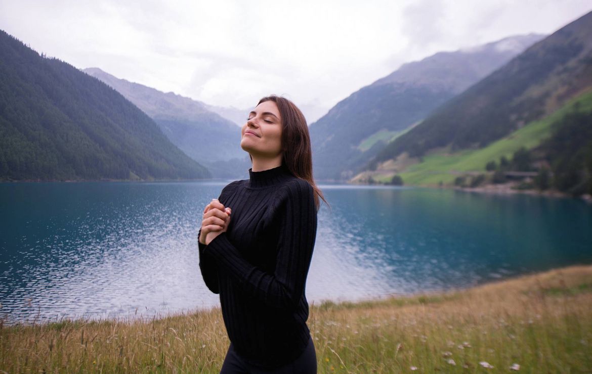 Eine Frau steht entspannt am Ufer eines Bergsees vor einer alpinen Landschaft.