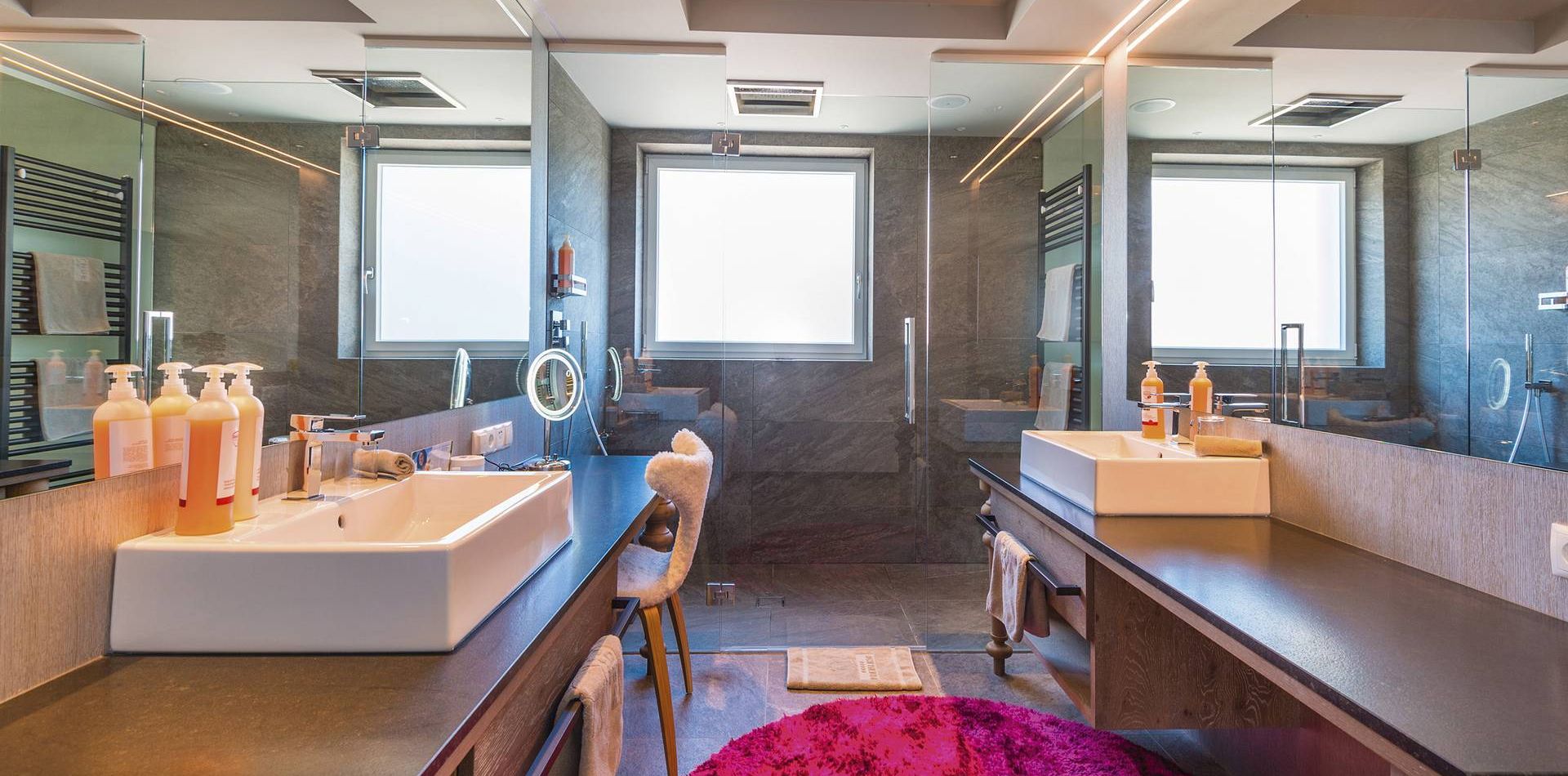 A modern suite bathroom with a double vanity, large mirror, walk-in shower and a bright window.