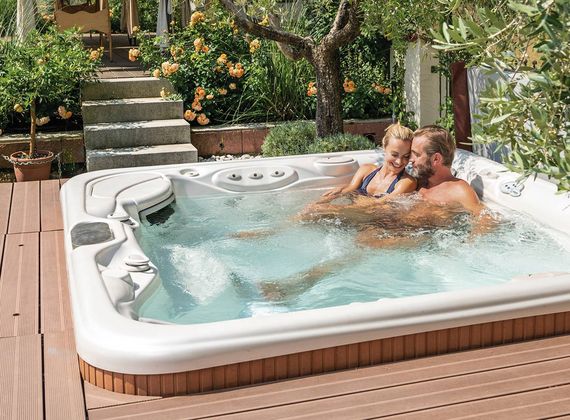 A couple relaxes together in an outdoor whirlpool surrounded by a wooden deck and garden greenery.