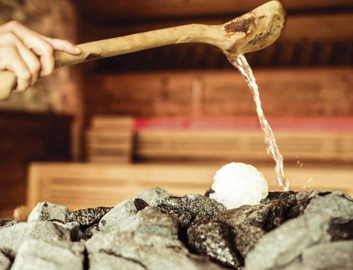 A hand pours water from a wooden ladle over sauna stones beside a pile of ice.