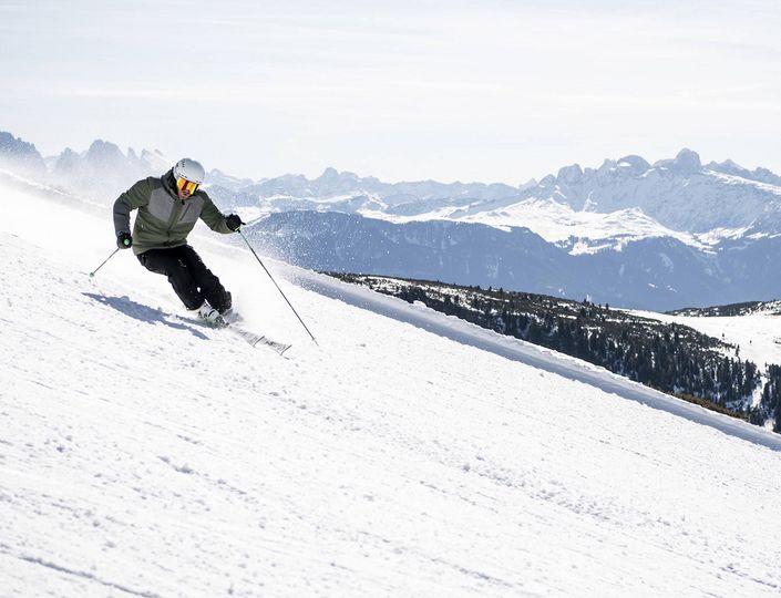 Ein Skifahrer fährt bei Sonnenschein über eine breite Piste mit Blick auf die verschneiten Berge.