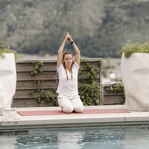 Eine Frau sitzt am Pool und macht Yoga zwischen zwei großen Pflanzgefäßen in Gesichtsform vor Bergkulisse.