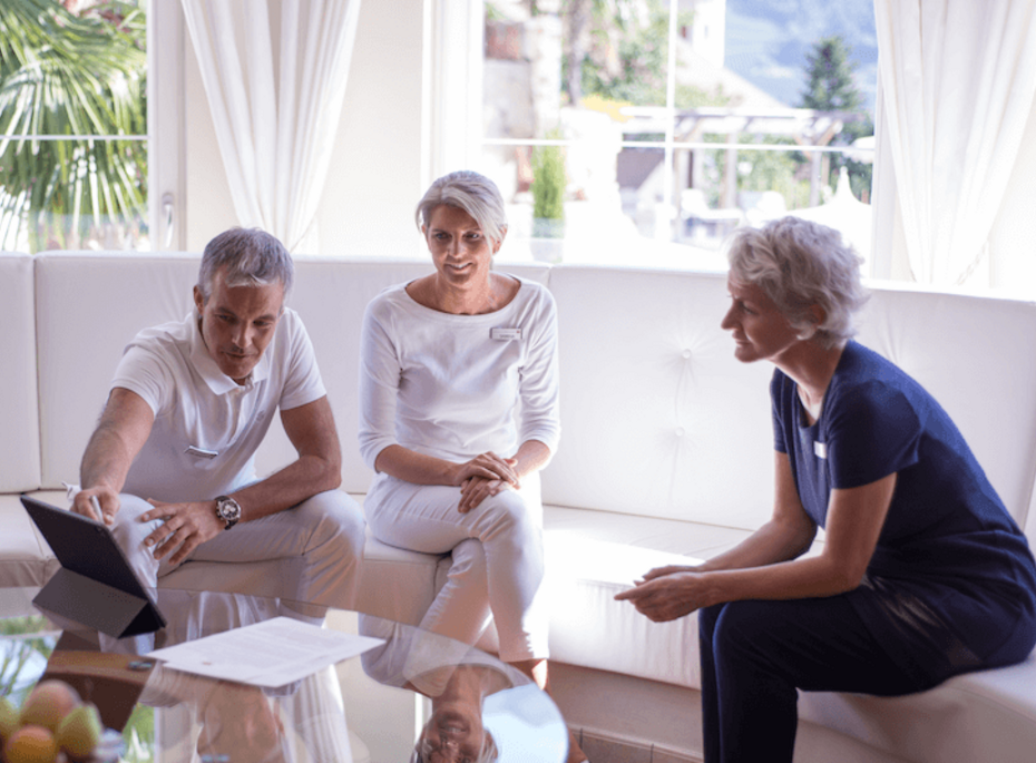 Wellnessberatung im Preidlhof in Naturns Drei Erwachsene sitzen in einer hellen Lounge und sprechen mit einer Beraterin am Glastisch.