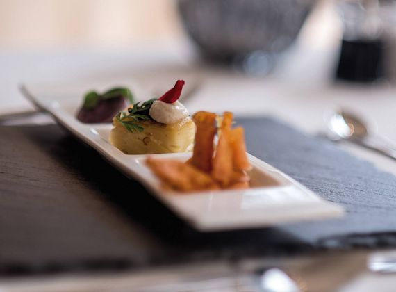 A plated gourmet dish is served on a rectangular white plate with a blurred dining room in the background.