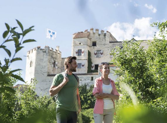 Ein Paar spaziert durch einen grünen Garten vor einem historischen Schloss mit wehender Fahne.