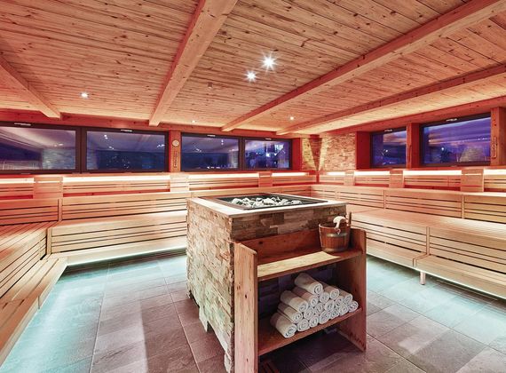 A wood-paneled sauna room with benches around a central heater and towels on the lower shelf.