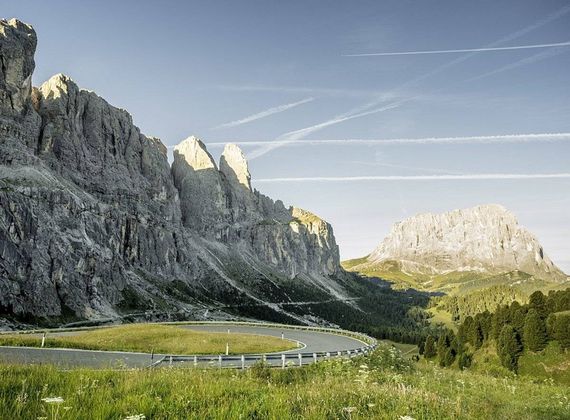 Una strada di montagna curva tra prati alpini sotto pareti rocciose ripide e un cielo limpido.