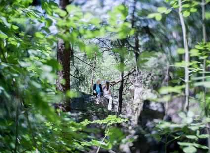 Zwei Personen sitzen auf einem Felsweg zwischen Bäumen und Grün im Wald.
