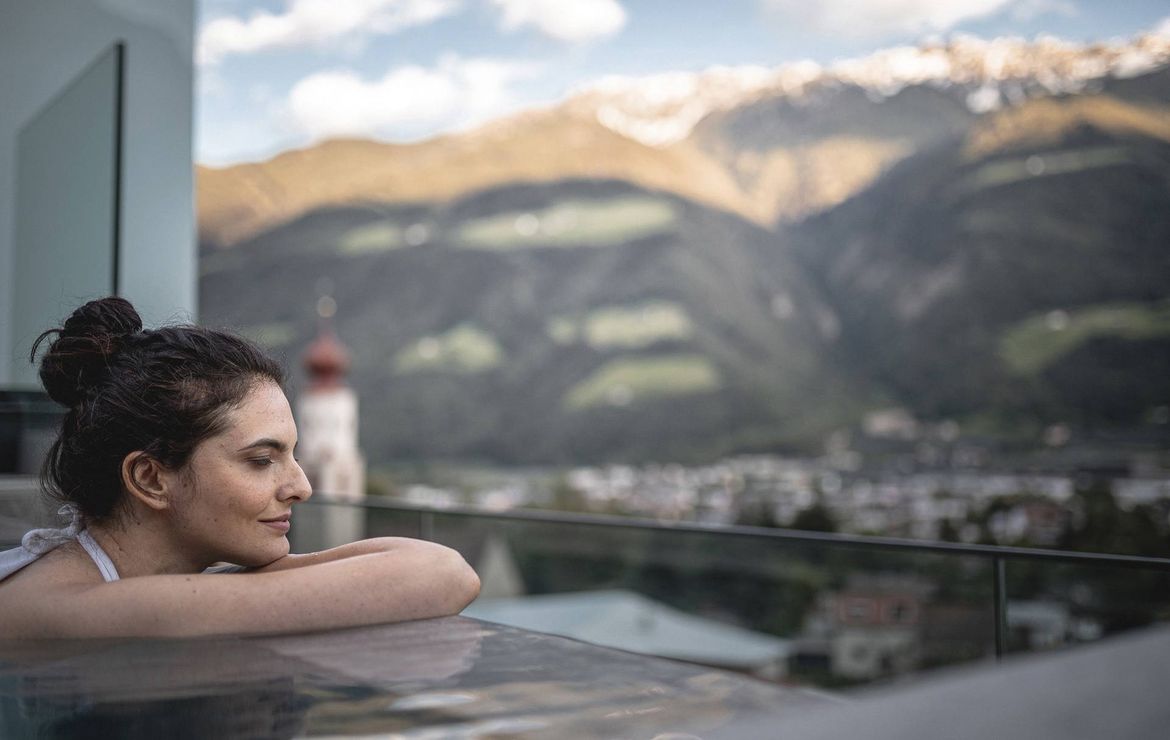 Una donna si rilassa in una piscina all'aperto con montagne e un paese sullo sfondo.