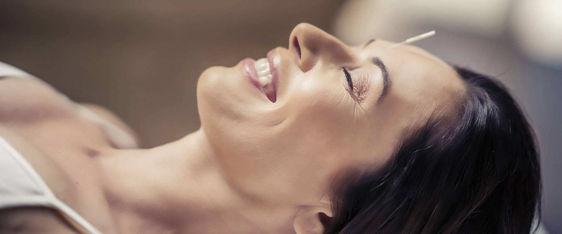 A smiling woman lies back during a relaxing spa treatment, with her face turned upward and eyes closed.