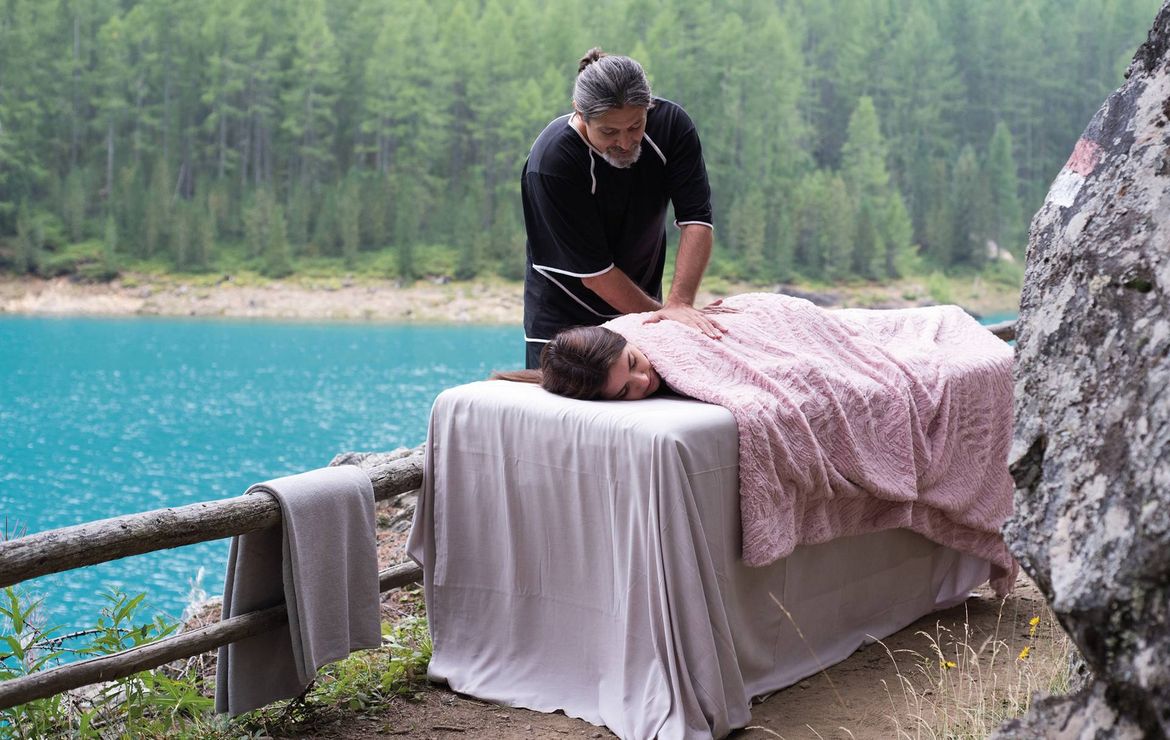 Ein Therapeut behandelt eine liegende Person auf einer Massageliege am türkisblauen Seeufer im Wald.