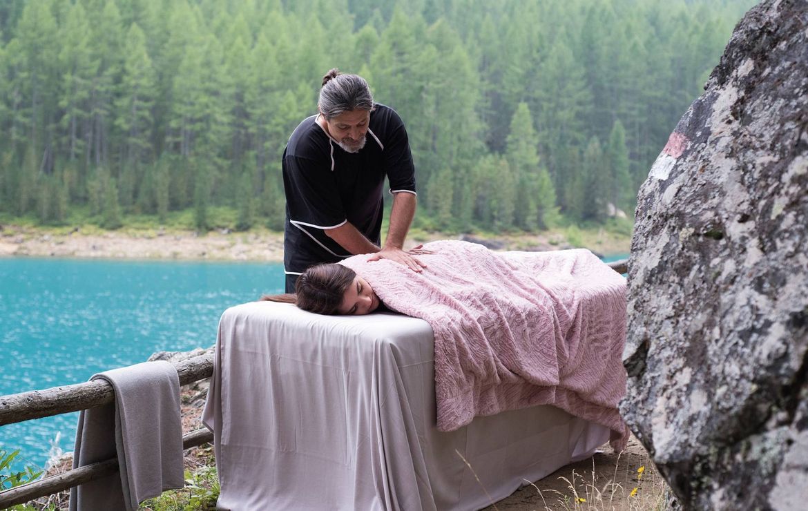 Un terapista esegue un massaggio all'aperto accanto a un lago turchese, con il cliente coperto su un lettino.