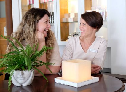 Zwei Mitarbeiterinnen unterhalten sich am Empfang neben einer Pflanze und einer beleuchteten Tischlampe.