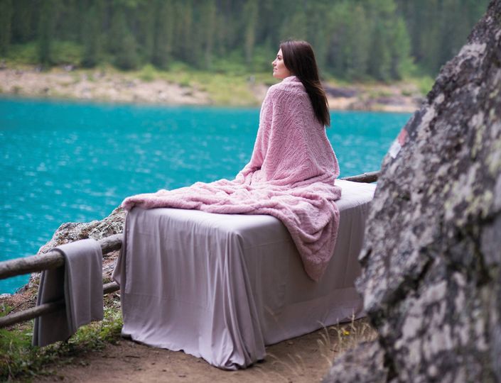 Eine Frau sitzt eingehüllt in eine rosa Decke auf einer Liege am türkisfarbenen See.