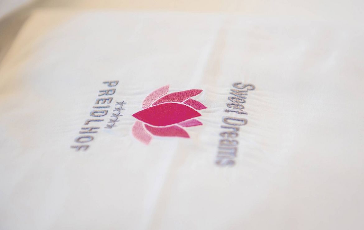 Ein Preidlhof-Logo mit pinker Blüte liegt auf hellem Stoff im Nahaufnahme-Ausschnitt.
