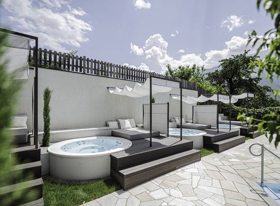 Eine Pool-Lounge mit zwei runden Wasserbecken, Liegen und Sonnensegeln auf einer Terrasse.