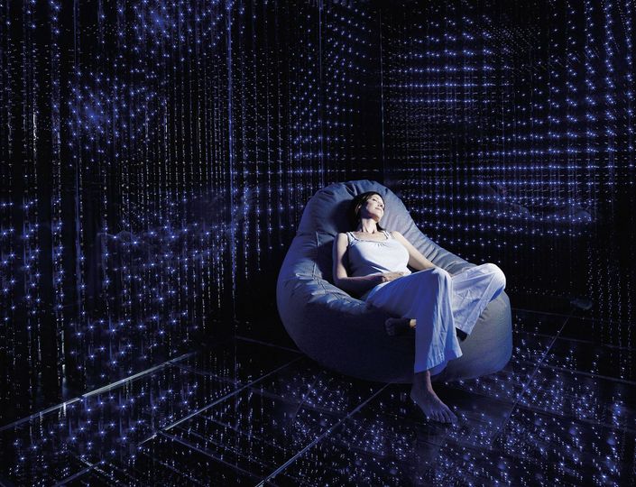 A woman reclines in a lounge chair inside a dark relaxation room with blue light points.