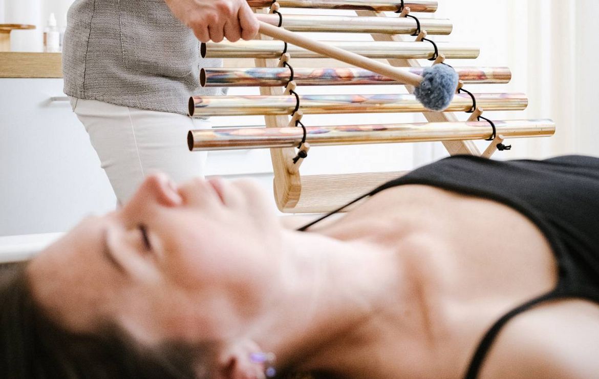 A woman lies on a treatment bed while a therapist plays tuning forks above her in a spa room.