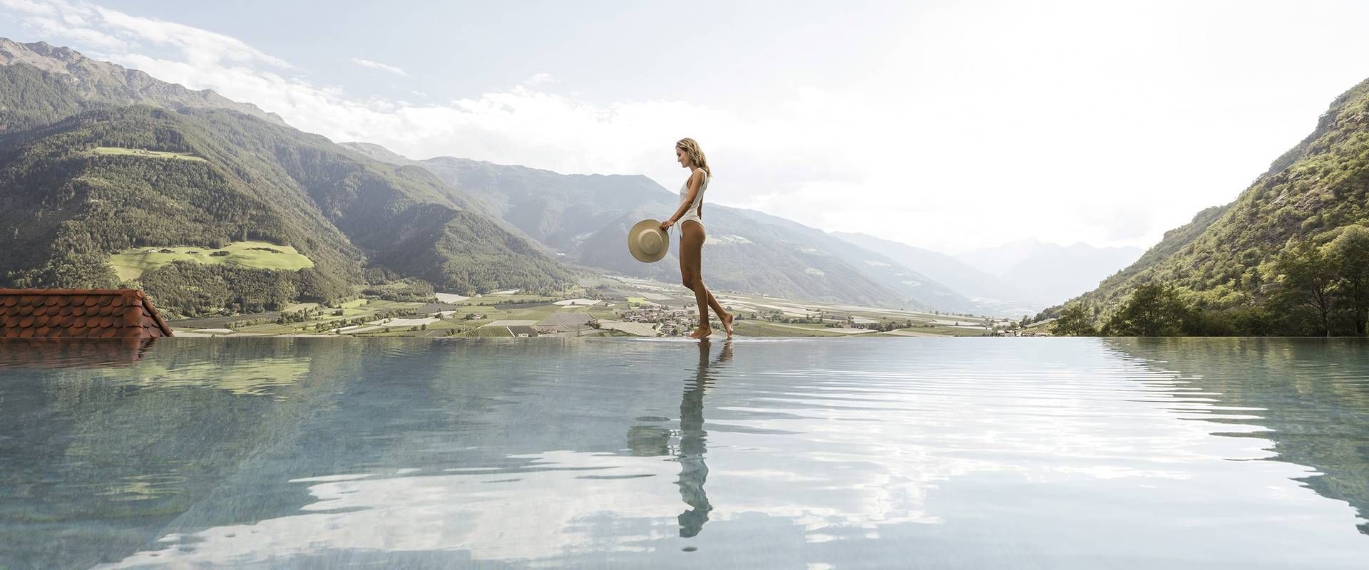 Eine Frau steht am Infinity-Pool vor einer alpinen Bergkulisse und hält einen Hut in der Hand.