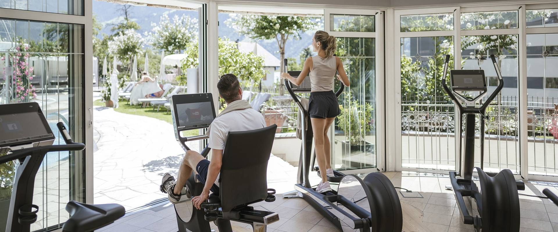 Un uomo usa una macchina per remare nella sala fitness mentre una donna è vicino alla porta vetrata aperta sul giardino.