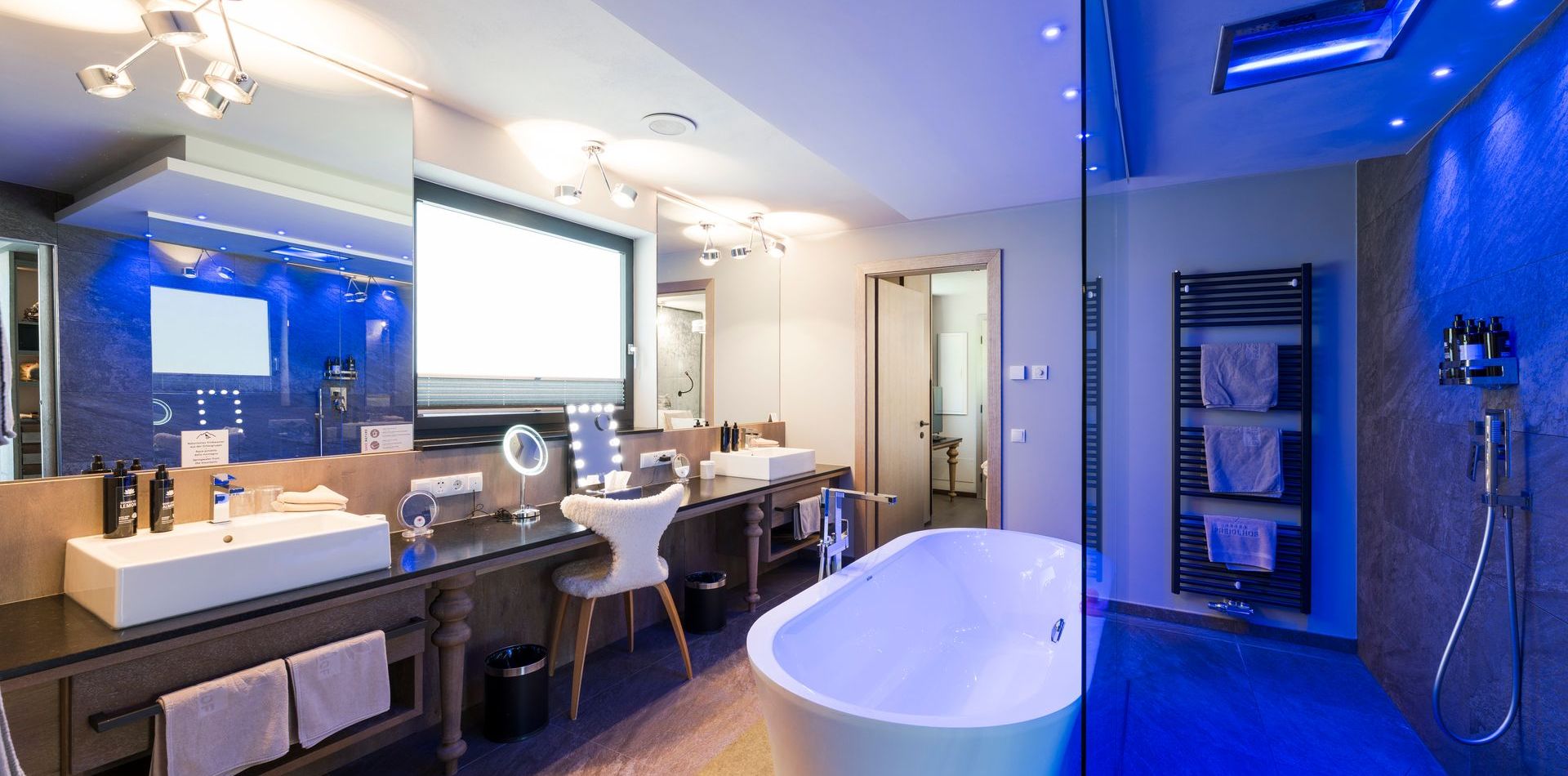 Badezimmer der Penthouse Suite mit freistehender Badewanne, Doppelwaschbecken, großem Spiegel und begehbarer Dusche mit blauer Beleuchtung.