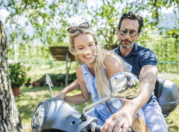 Ein Paar fährt gemeinsam auf einer Vespa im Grünen und lächelt in die Kamera.