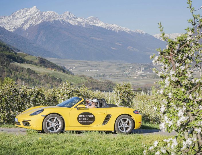 A yellow convertible with DolceVita branding is parked in front of South Tyrolean mountains and orchards.